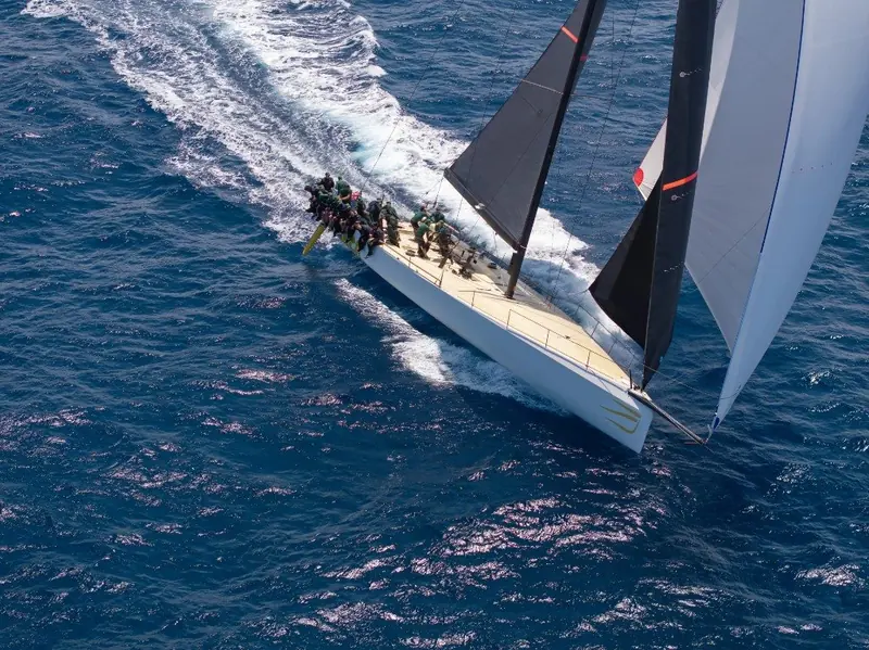 Close-hauled with genoa and mainsail trimmed tight, crew working the rail as the bow drives through a wave.