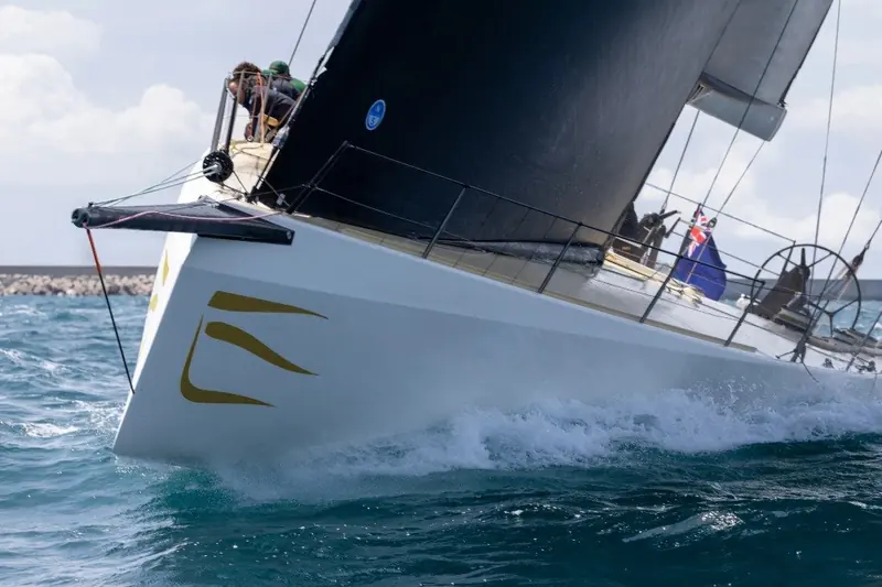 Bow pulpit and forestay detail as the crew trims through a tack, black carbon mast and standing rigging visible against the sky.