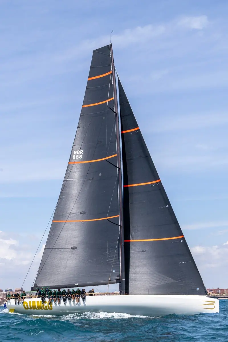 Full rig upwind under mainsail and headsail, the carbon mast raked aft and the orange accent stripe running the length of the hull.