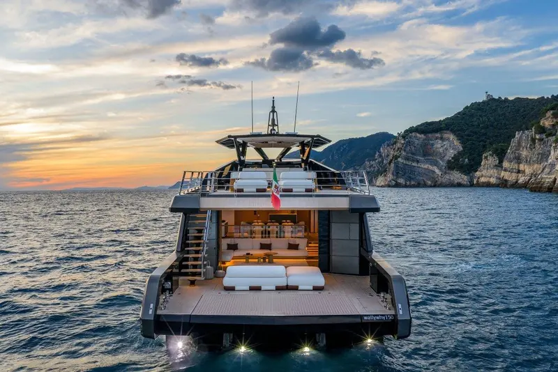 The wallywhy150's transom at dusk off the Ligurian coast, name badge lit on the swim platform and the aft cockpit lounge glowing warm against the sunset.