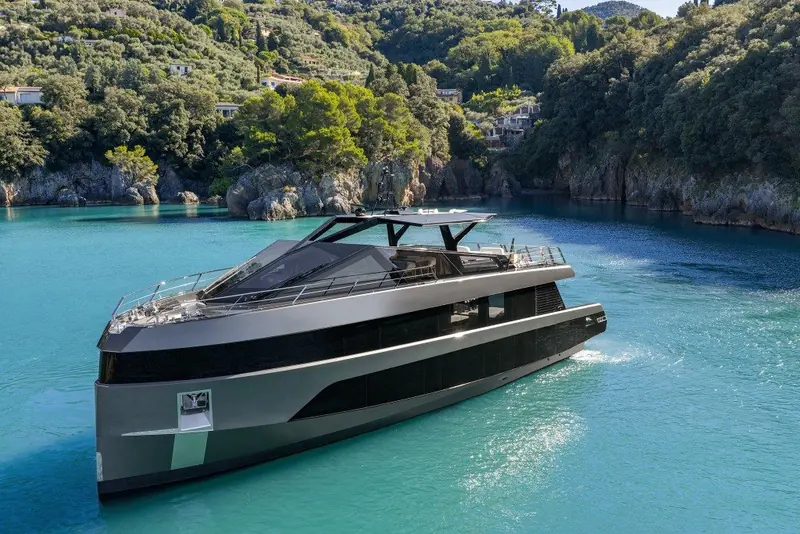 Three-quarter view at anchor in a turquoise bay, terraced hillside villas and umbrella pines climbing the shore behind.