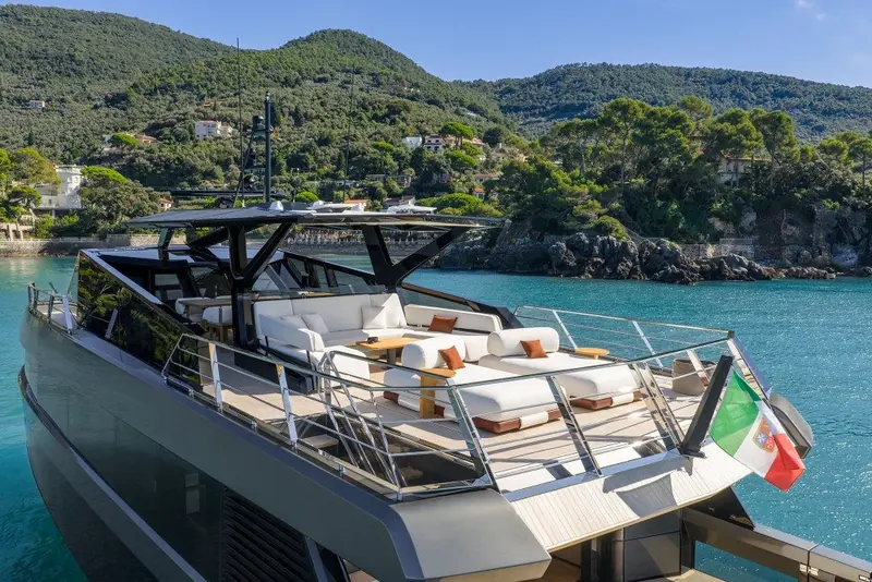 The flybridge lounge seen from the foredeck, white cushions and a teak table under the open hardtop, Italian ensign flying off the transom.