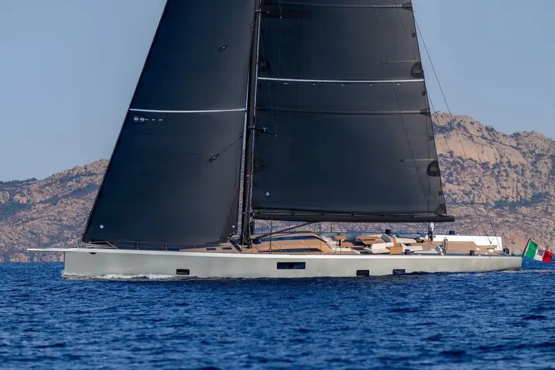 Sailing close to a rocky coastline under black sails, the hull's low freeboard keeping her profile lean against the hills behind.