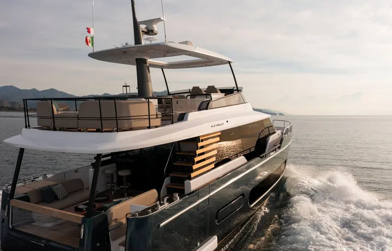 Stern view under way showing the MAGELLANO 60 nameplate, the teak boarding stair folded against the transom, and the flybridge seating above.