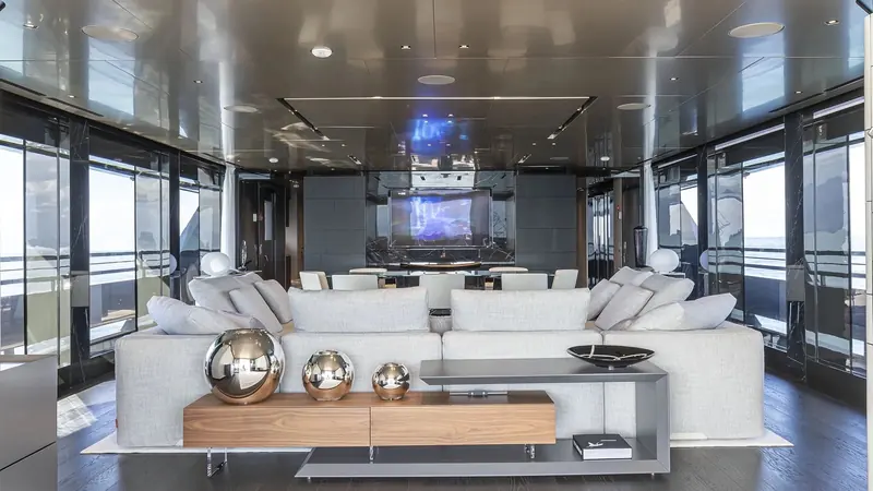 The main saloon, dark lacquer and marble surfaces against full-height glazing, with the dining table set behind the sofa.