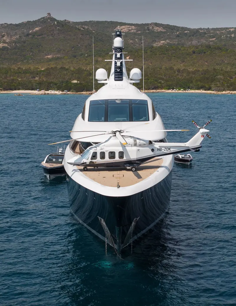 A Swiss-registered AW139 parked on the foredeck helipad off Corsica, with the yacht's own tenders standing off on each beam.