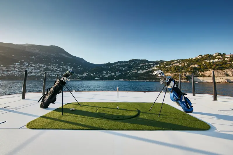 A full golf setup with mats and bags laid out on the foredeck, Monaco's hillside visible across the bay.