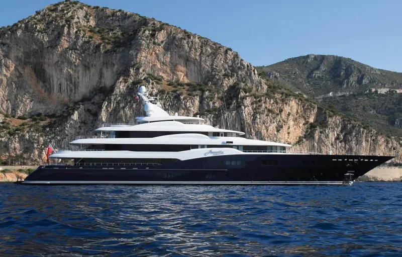 Amaryllis against limestone cliffs on the Mediterranean coast, her hull colour matching the deep water beneath her.