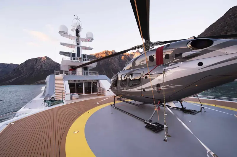 The helicopter secured on its dedicated foredeck pad, mountains and fjord ice visible beyond the rail - Cloudbreak carries a helicopter as standard equipment for a boat this size, not an occasional charter add-on.