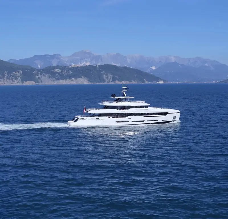 Under way against a hazy mountain coastline, the low, level running attitude typical of Baglietto's displacement-leaning hulls rather than the bow-up stance of a planing yacht at speed.