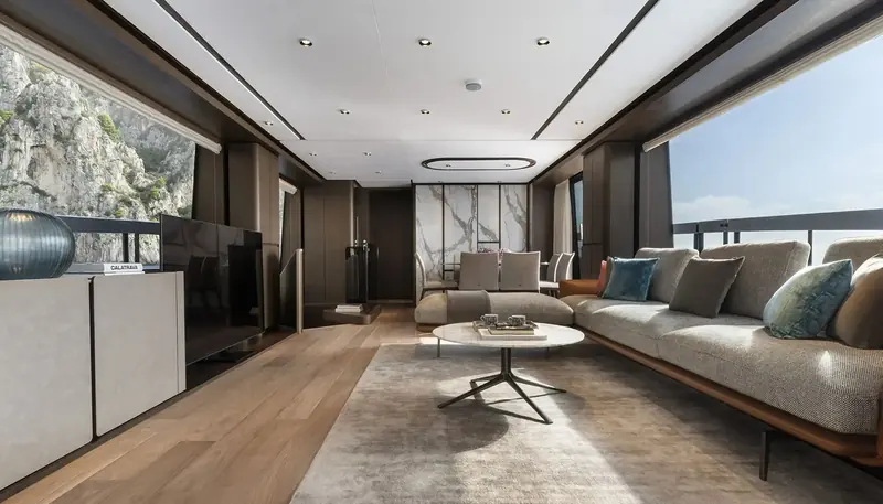 The Classico-scheme main saloon pairs pale oak flooring with a bookmatched marble panel and a sectional sofa, with full-height glazing framing the cliffs outside.