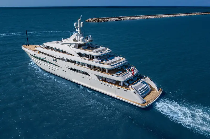 An aerial view of Mimtee arriving past a breakwater, guests gathered on the aft deck beside the raised tender-garage door as she slows toward harbour.