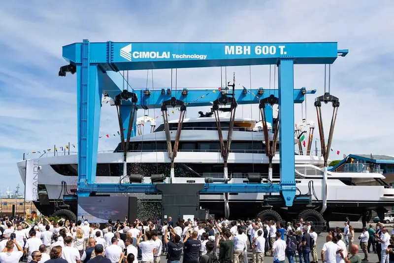 The 600-tonne Cimolai travel lift carries the hull toward the water as the launch-day crowd gathers beneath it.
