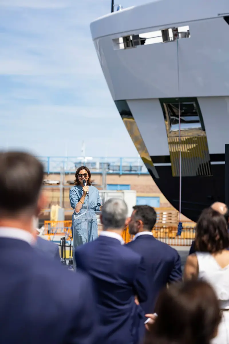 A speaker addresses the naming-ceremony crowd from beside the hull.