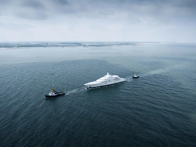 Two tugs escort Project Tanzanite off the Dutch coast near Vlissingen, the low profile of the Westerschelde barrier islands behind her.