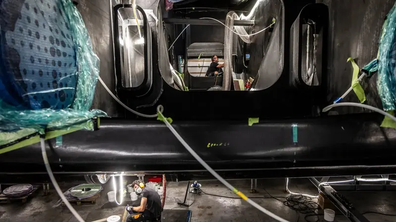 Inside the build shed: a technician works on Hypersail's structure beneath a hull section, with the resin, ducting and protective wrapping of an active composite yard around them.