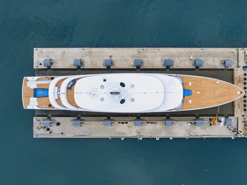 A drone's view straight down onto Belita at the quay shows the full 12.5-metre beam and the foredeck layout before her main-deck fit-out is finished.