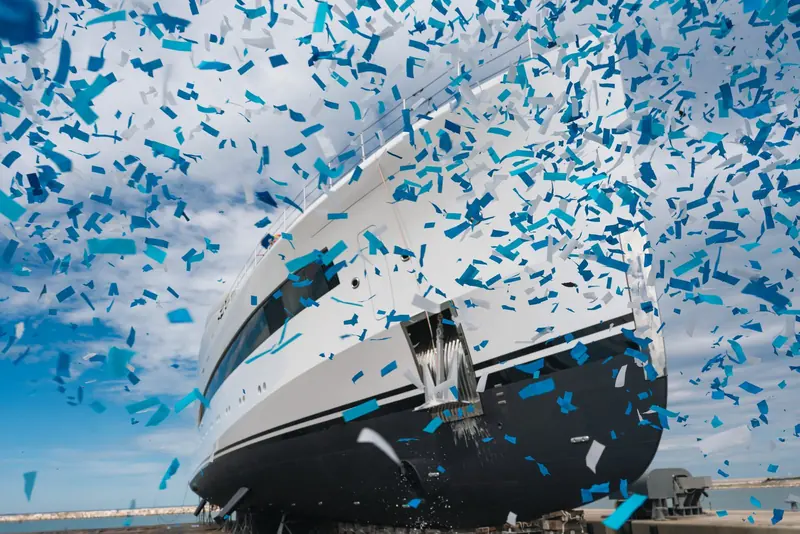 Blue and white confetti falls across Belita's bow at the moment of naming - the hull's black boot-top and anchor pocket visible beneath.