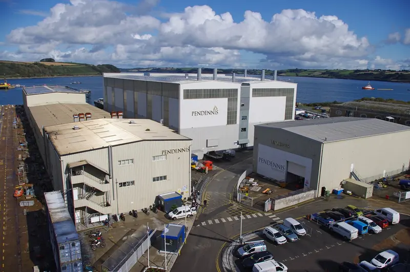 Pendennis at Falmouth Docks, with the Carrick Roads behind. The tall white shed is one of the two 90-metre construction halls; the yard's whole Falmouth operation sits on 14 acres of this waterfront, which is why the land next door matters.