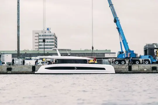 The travel crane lowers Pioneer One toward the water, her low, single-level profile visible against the yard buildings.