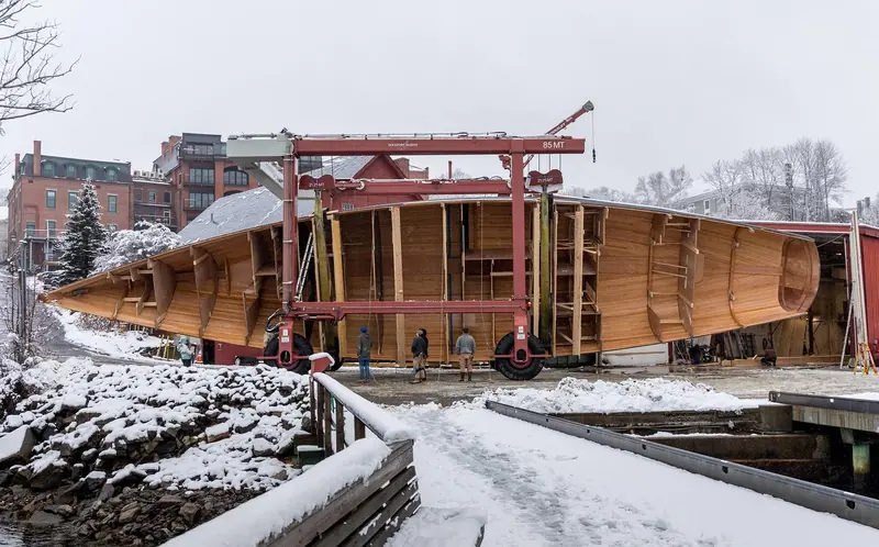 The finished hull, built in strip-planked wood, moves on Rockport Marine's travel lift past the yard's Maine waterfront in winter snow before fitting out.