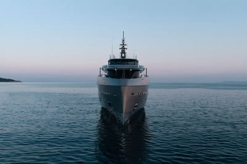 Heed approaches head-on at dusk, her metallic grey hull and raked bow reflected in a calm sea.