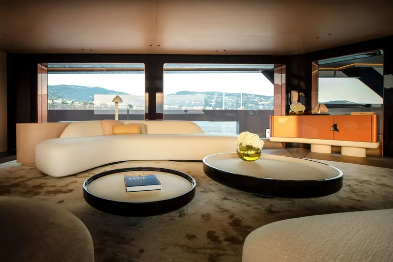 The main saloon, with curved cream seating, a lacquered orange sideboard and wide windows onto the coast.