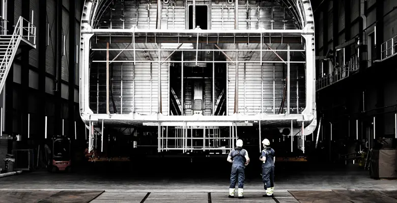Two shipyard workers look up at the open aluminium framing from the floor of the hall, giving a sense of scale that no rendering can.