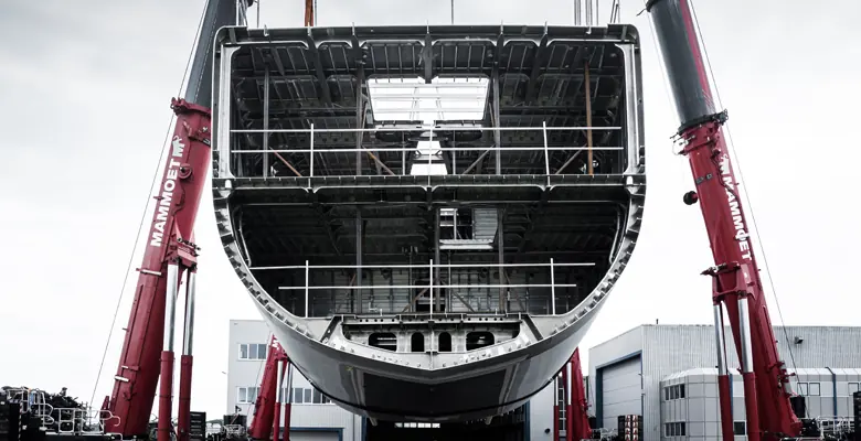 The stern framed by the cranes at the moment of the turn, the hull's raw aluminium skin still unpainted.