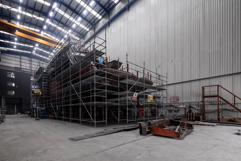 Scaffolding wraps the full length of the hull inside the build hall, framed against the shed's roof trusses.