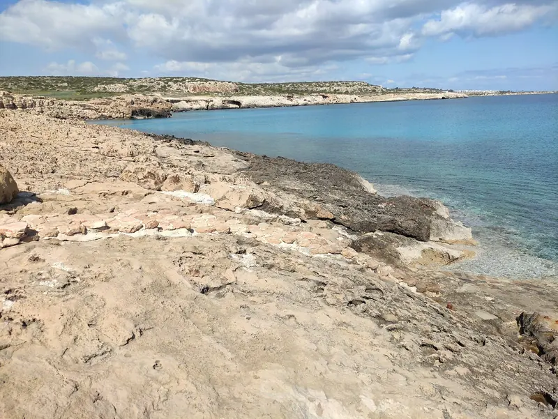 The low, rocky shoreline typical of Cape Greco, the kind of lee shore a boat is driven onto when wind and swell turn against an open anchorage.