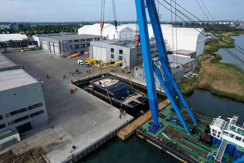 A wider view of the launch basin: the crane barge, the covered sheds and the hull suspended over open water.