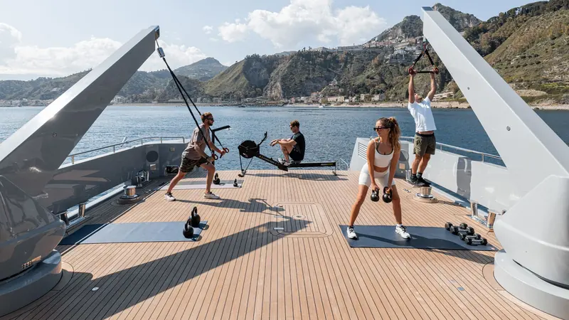 Guests exercise in the open-air gym rigged on the foredeck, with a hillside town on the coast behind.