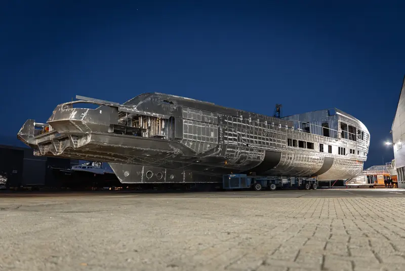 Project Zen's joined hull and superstructure stands on a heavy transporter in the Waalwijk yard after dark, the aluminium framing and window openings still bare.