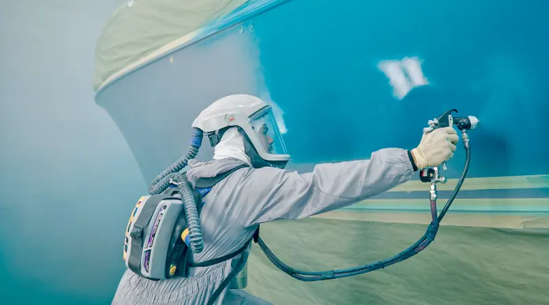 An applicator spray-applies Awlgrip topcoat to a Hinckley hull inside a controlled paint booth.