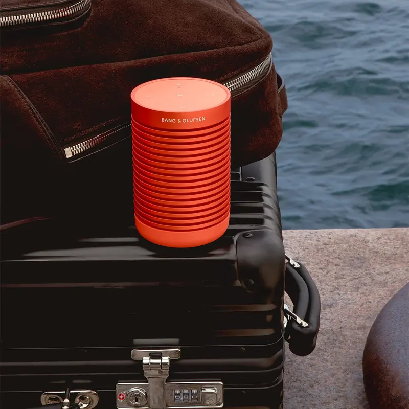 A Beosound Explore in Bonfire Orange clipped to a Rimowa case at the waterside - the self-contained end of the catalogue, next to the integrator-installed systems that reach yachts.