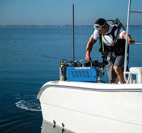 A diver suits up on a tender's swim platform beside a blue portable compressor, mask and regulator already on, one hand resting on the unit's control panel.
