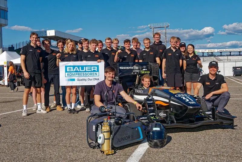 A Bauer JUNIOR II portable compressor stands trackside next to an open-wheel racing car, its team crouched around a sponsorship banner at Hockenheimring.