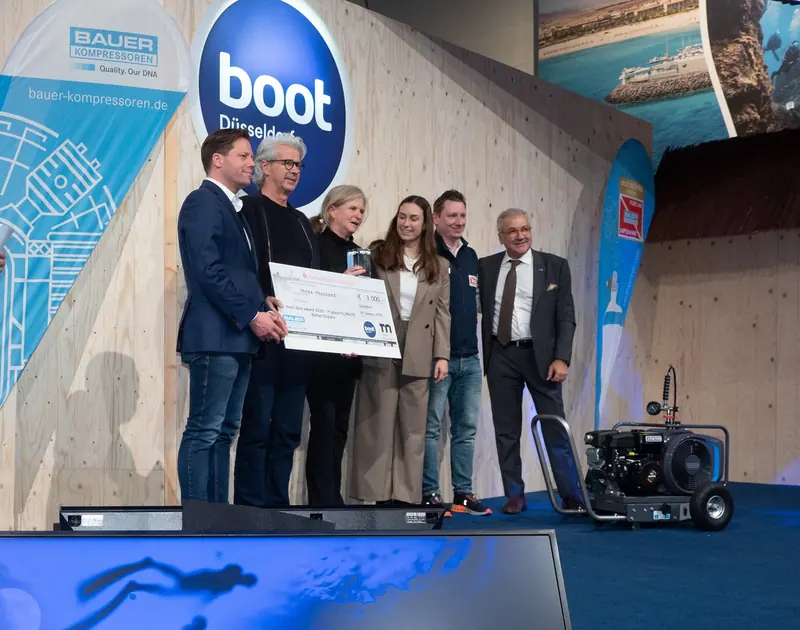 A dive-award ceremony at the boot Dusseldorf trade show, winners holding an oversized cheque on stage beside a Bauer portable compressor set up for the demonstration.