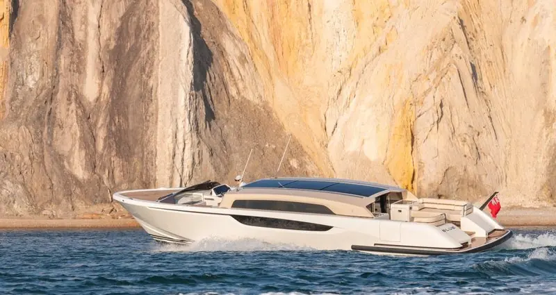 Sunlit rock rises behind a cream limousine tender making way close inshore.