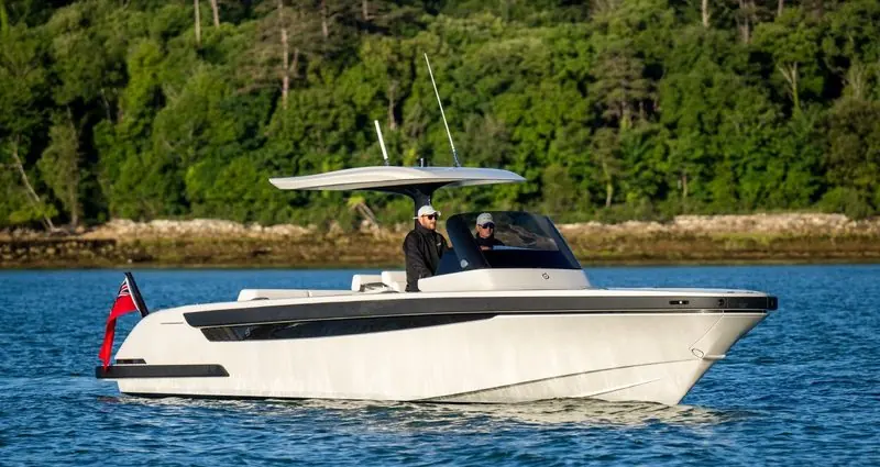 Under a low hardtop, two people take a tender with a dark boot stripe along a wooded shore.
