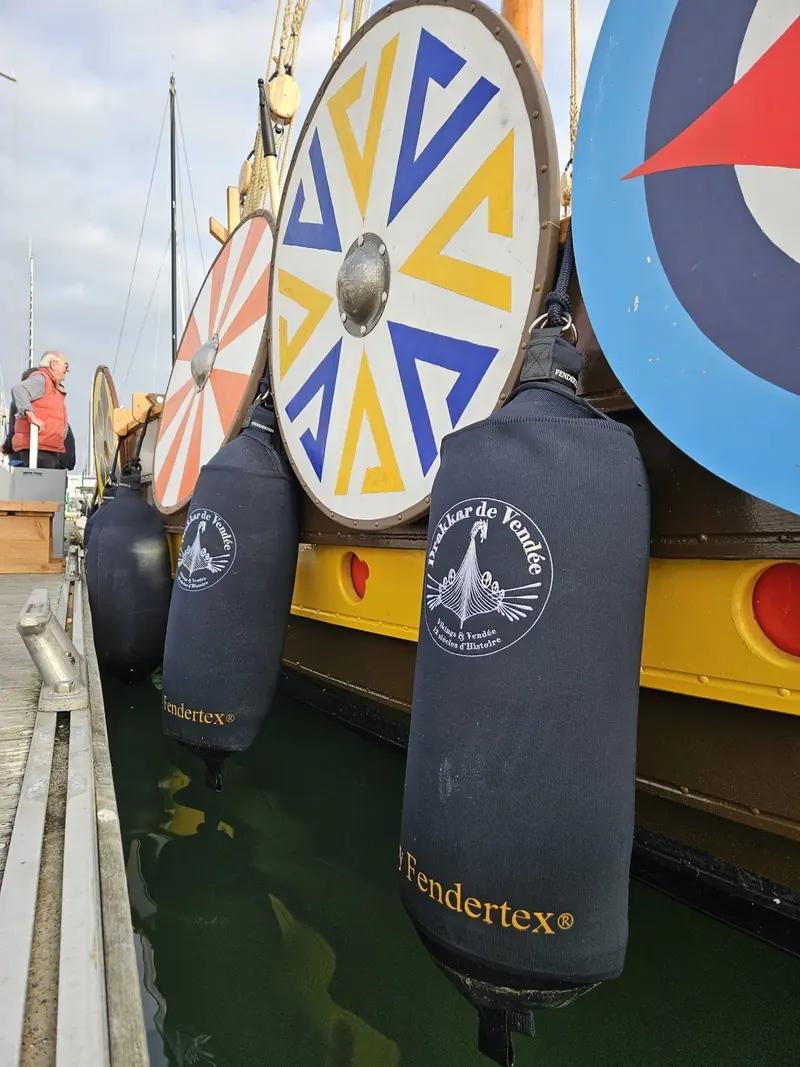 A close-up of two Fendertex cylindrical fenders custom-embroidered with a client's own crest and yellow Fendertex branding on the woven cover, hung beside painted Viking-style shields on a replica longship in Vendee.