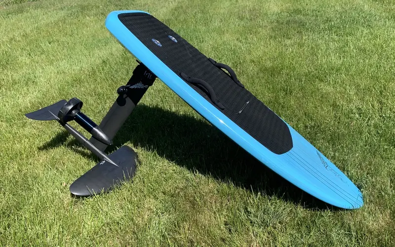 The complete board propped upright on its mast, front wing planted in the grass, showing the padded deck strap and twin footwell traction pads that keep a rider locked on through a hard turn.