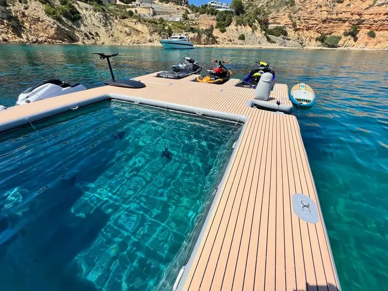 Close alongside, a floating teak-topped dock encloses a netted swimming pool with jet skis parked on the far side.