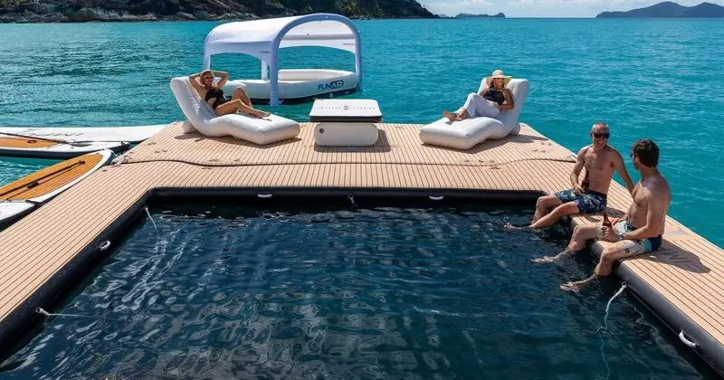 Guests sit on the edge of a floating dock beside a shaded daybed, feet over the netted pool.