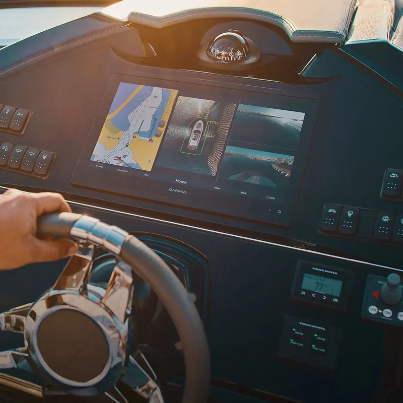 A helmsman's hand on the wheel of a motoryacht, a Garmin multifunction chartplotter on the dash split into chart, surround-view camera and sonar panes, Volvo Penta engine displays mounted just below it.