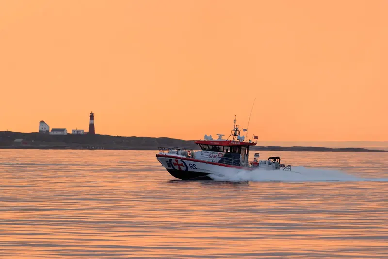 A Norwegian Redningsselskapet search and rescue boat running at speed past a lighthouse at dusk, the kind of high-speed commercial hull where interceptor trim control has to work in a following sea, not just at the dock.