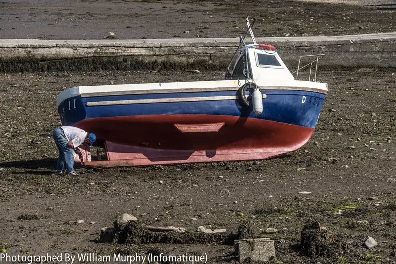 A small workboat hauled out at Malahide, Ireland, showing a fresh red antifouling bottom coat against dark blue topsides while its owner works on the propeller.