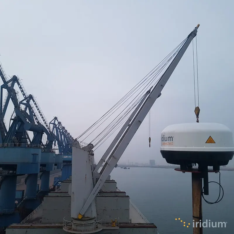 An Iridium Certus maritime broadband antenna being hoisted onto a vessel at a container port; the low-profile radome holds the phased-array antenna used by Certus 100, 200 and 700 terminals.