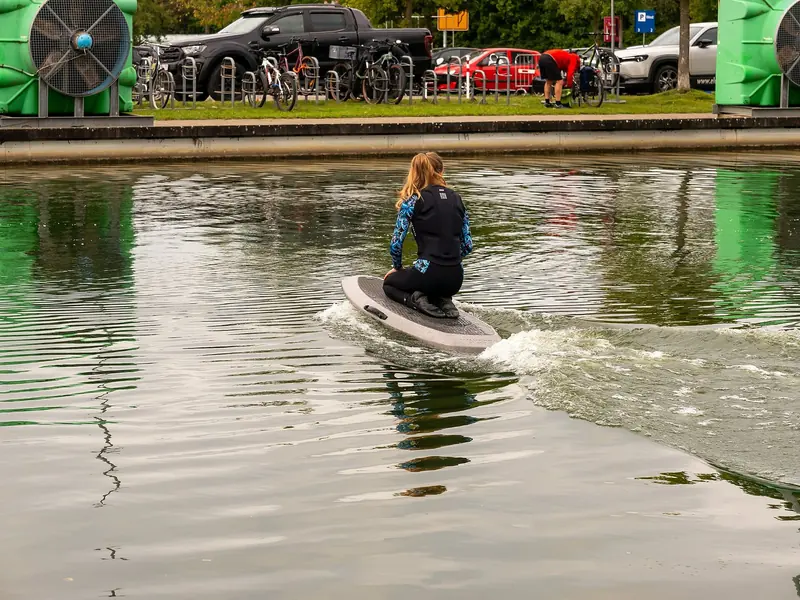 A rider kneels low over the board on a calm show-lake circuit, spectators' bikes racked on the bank behind her.